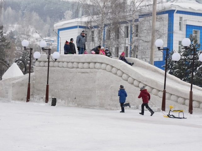 Children are playing on a large snow slide in a winter setting. Some kids are sitting on top, while others are running nearby. The scene includes multiple lamp posts with spherical lights, and a building with blue and white walls in the background. Snow covers the ground and trees, creating a festive and playful atmosphere.