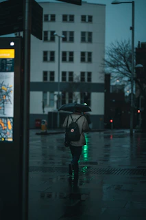 A traveler wearing a grey backpack walking through a bustling city street at dusk.