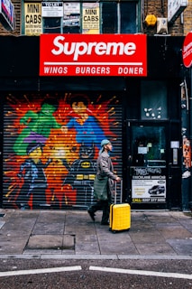 A person in a coat and hat walks on a city sidewalk, pulling a bright yellow suitcase. Behind them is a storefront featuring colorful graffiti of superhero characters including the Hulk, Superman, and Batman painted on a metal shutter. Above the shutter, a red and white sign reads 'Supreme Wings Burgers Doner'. Various signage related to minicab services is visible, along with text and contact numbers.