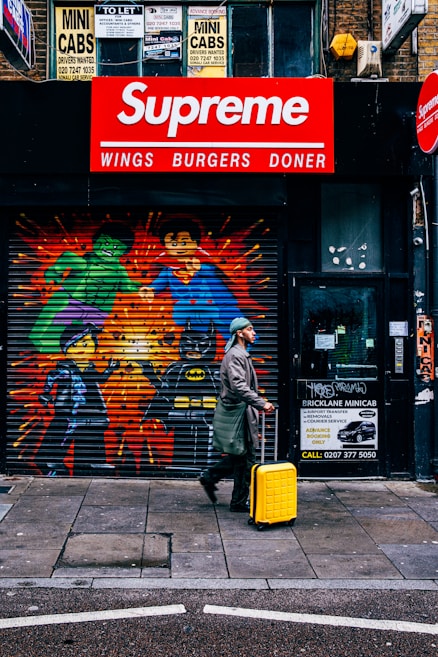 A person in a coat and hat walks on a city sidewalk, pulling a bright yellow suitcase. Behind them is a storefront featuring colorful graffiti of superhero characters including the Hulk, Superman, and Batman painted on a metal shutter. Above the shutter, a red and white sign reads 'Supreme Wings Burgers Doner'. Various signage related to minicab services is visible, along with text and contact numbers.