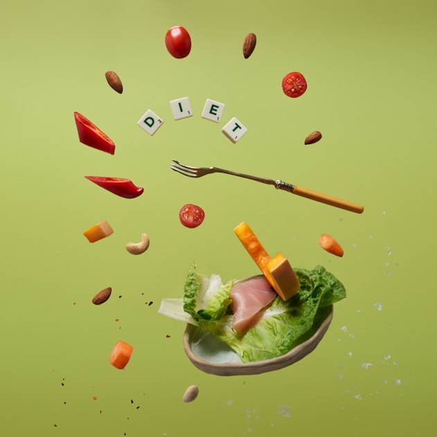 Floating food items including cherry tomatoes, almonds, cashews, slices of red bell pepper, cheese cubes, lettuce, and ham. A fork is levitating among these ingredients. The letters spelling 'DIET' are arranged with small tiles like a game. The background is a solid light green color.