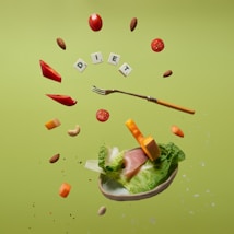 Floating food items including cherry tomatoes, almonds, cashews, slices of red bell pepper, cheese cubes, lettuce, and ham. A fork is levitating among these ingredients. The letters spelling 'DIET' are arranged with small tiles like a game. The background is a solid light green color.