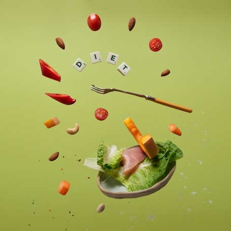 Floating food items including cherry tomatoes, almonds, cashews, slices of red bell pepper, cheese cubes, lettuce, and ham. A fork is levitating among these ingredients. The letters spelling 'DIET' are arranged with small tiles like a game. The background is a solid light green color.