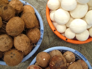 Three baskets are filled with coconuts, with the top basket containing peeled white coconuts, the bottom left holding whole coconuts with husks, and the bottom right containing partially peeled coconuts. The baskets are placed on a gray stone surface.