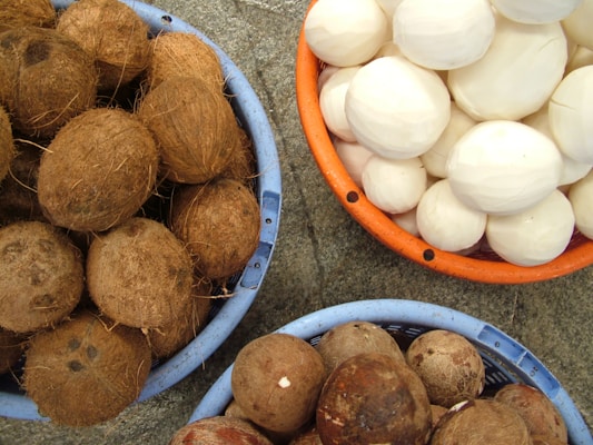 Three baskets are filled with coconuts, with the top basket containing peeled white coconuts, the bottom left holding whole coconuts with husks, and the bottom right containing partially peeled coconuts. The baskets are placed on a gray stone surface.