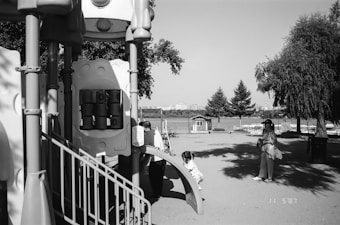 A playground with climbing structures made from sturdy materials is prominently featured. In the foreground, a child is climbing a small ramp. A woman stands nearby, looking on attentively, holding some belongings. Trees and a view of a body of water with buildings in the distance occupy the background, evoking a serene and open environment.