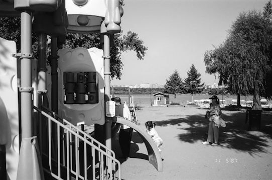A playground with climbing structures made from sturdy materials is prominently featured. In the foreground, a child is climbing a small ramp. A woman stands nearby, looking on attentively, holding some belongings. Trees and a view of a body of water with buildings in the distance occupy the background, evoking a serene and open environment.