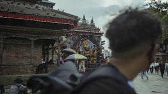 A historical temple site features intricate architecture with a prominent statue of a deity in the center, surrounded by people walking by and pigeons perched on the roof. An individual in the foreground looks towards the temple, while several pedestrians and a black goat are visible near the structure. The atmosphere appears overcast with clouds in the sky.