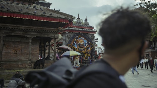 A historical temple site features intricate architecture with a prominent statue of a deity in the center, surrounded by people walking by and pigeons perched on the roof. An individual in the foreground looks towards the temple, while several pedestrians and a black goat are visible near the structure. The atmosphere appears overcast with clouds in the sky.