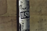 A street pole covered in various black and white posters, prominently featuring a poster with the text 'Black Lives Matter' alongside an image of a raised fist. The background is a blurred stone wall, creating an urban setting.