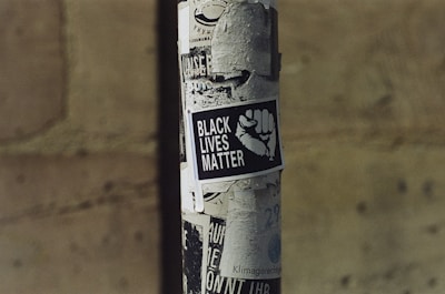 A street pole covered in various black and white posters, prominently featuring a poster with the text 'Black Lives Matter' alongside an image of a raised fist. The background is a blurred stone wall, creating an urban setting.