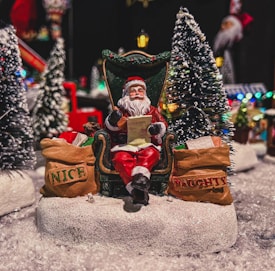 A festive scene featuring a Santa Claus figurine sitting on a green throne-like chair. Santa is holding a list, with bags labeled 'NICE' and 'NAUGHTY' on either side, surrounded by snow and miniature pine trees.