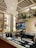 A cozy bar setting is adorned with hanging plants and lights, featuring a well-organized counter displaying glasses and beverages. Shelves contain an array of bottles and decorations, creating a welcoming ambiance. The ceiling has a patterned design that enhances the overall decor.