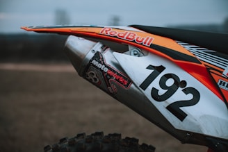 A close-up view of a motorcycle exhaust pipe with a Red Bull sticker and the number 192 displayed prominently. The bike features an orange, white, and black color scheme with additional decals, including one with the word 'Motoworks'. The background is blurred, suggesting an outdoor setting.