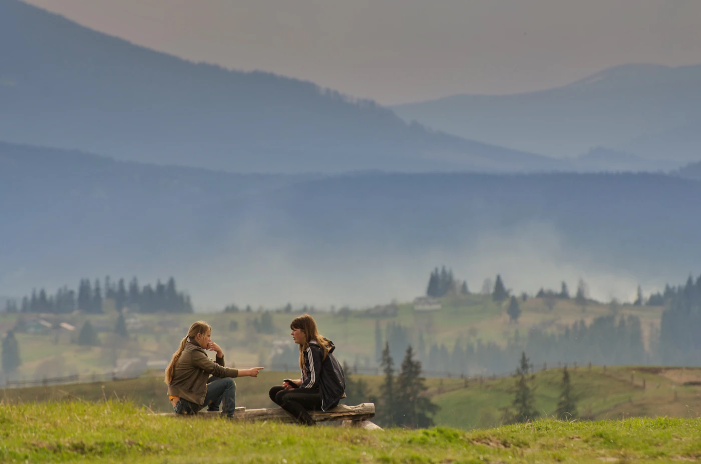 Two hikers paused on a high grassy ridge in the Carpathians, turned toward each other mid-conversation—epic layered peaks and big-sky trail country behind them