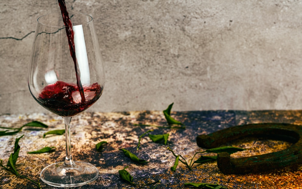 A glass of wine is being poured with a background featuring a rustic table scattered with green leaves and a rusted horseshoe. The scene conveys a sense of rustic charm and casual elegance.