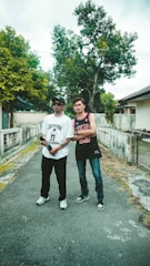 Two young individuals are standing on a narrow pathway in an outdoor setting. One is wearing a white graphic T-shirt, black jeans, sunglasses, and a hat, while holding a bottle. The other is dressed in a black basketball jersey, blue jeans, and sneakers. They appear relaxed and casually pose for the photo. Tall trees with green leaves and residential buildings are visible in the background.