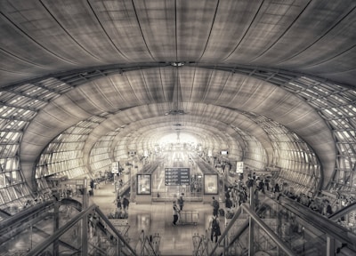 A spacious, futuristic airport terminal with a high arched ceiling made of large, evenly spaced panels. Numerous people are present, engaging with various airport facilities such as information desks and security checkpoints. The environment is illuminated with a combination of natural and artificial light, emphasizing the grand architecture.