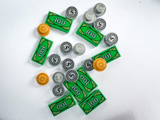 A collection of toy currency items scattered on a white surface, including green rectangular bills marked with 100 and round coins marked with 5 and 1. There are also a few orange and gray circular pieces.