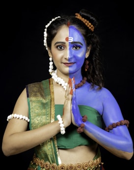 A woman is standing with her hands joined in a traditional greeting pose. Her face and upper body are painted to represent a half-and-half imagery, combining feminine features with symbolic elements. Her left side is painted in blue with adornments that resemble traditional religious or mythological attire. She wears jewelry and a green and gold saree.
