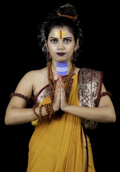 A person dressed in a traditional Indian outfit with a yellow saree adorned with intricate designs. They have several rudraksha beads around their neck and arms. The person is holding their hands in a Namaste gesture, with decorated forehead markings and intense facial expression.