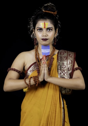 A person dressed in a traditional Indian outfit with a yellow saree adorned with intricate designs. They have several rudraksha beads around their neck and arms. The person is holding their hands in a Namaste gesture, with decorated forehead markings and intense facial expression.