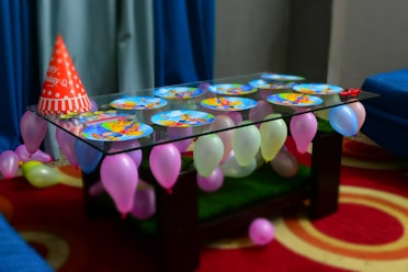 A glass table is adorned with colorful, cartoon-themed paper plates and surrounded by vibrant balloons in various pastel colors. A red 'Happy Birthday' party hat is placed on the table along with a small red toy car. The table is set against a backdrop of blue curtains and is placed on a red carpet with circular patterns.