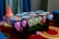 A glass table is adorned with colorful, cartoon-themed paper plates and surrounded by vibrant balloons in various pastel colors. A red 'Happy Birthday' party hat is placed on the table along with a small red toy car. The table is set against a backdrop of blue curtains and is placed on a red carpet with circular patterns.