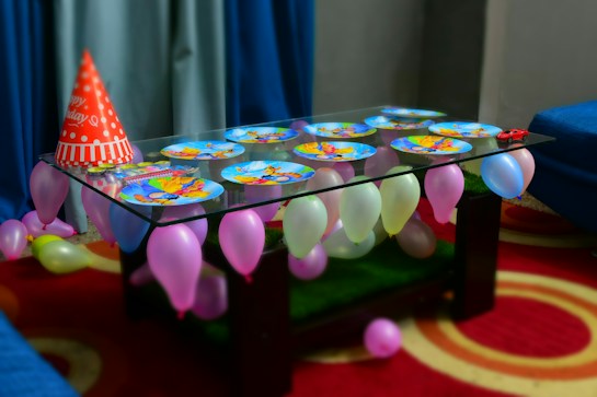 A glass table is adorned with colorful, cartoon-themed paper plates and surrounded by vibrant balloons in various pastel colors. A red 'Happy Birthday' party hat is placed on the table along with a small red toy car. The table is set against a backdrop of blue curtains and is placed on a red carpet with circular patterns.