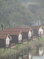 Several wooden cabins with red roofs are elevated on stilts above a calm body of water, reflecting their images. The cabins are aligned in a row, facing the water. Surrounding the area are trees with sparse foliage, indicating a colder season or early spring. The overall setting appears to be tranquil and natural.