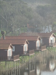Several wooden cabins with red roofs are elevated on stilts above a calm body of water, reflecting their images. The cabins are aligned in a row, facing the water. Surrounding the area are trees with sparse foliage, indicating a colder season or early spring. The overall setting appears to be tranquil and natural.