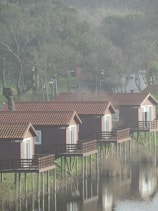 Several wooden cabins with red roofs are elevated on stilts above a calm body of water, reflecting their images. The cabins are aligned in a row, facing the water. Surrounding the area are trees with sparse foliage, indicating a colder season or early spring. The overall setting appears to be tranquil and natural.