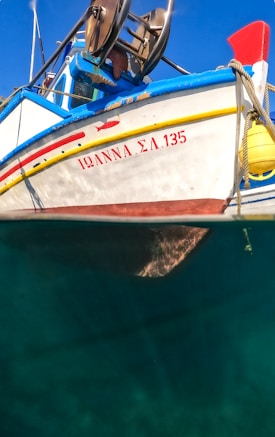 A boat with a blue and white hull is partially submerged in clear water. The name 'ΙΩΑΝΝΑ ΣΛ 135' is written in red on the side. Yellow and red stripes add color to the design, and there are visible parts of fishing equipment on board. The background shows a bright blue sky.