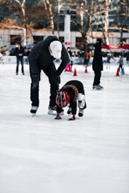 A person wearing a white cap and black clothing is helping a child in a red helmet who appears to be learning how to ice skate. The scene takes place on an ice rink with other skaters in the background and trees with no leaves. Orange cones are set up on the ice, possibly for guidance or lessons.