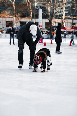 A person wearing a white cap and black clothing is helping a child in a red helmet who appears to be learning how to ice skate. The scene takes place on an ice rink with other skaters in the background and trees with no leaves. Orange cones are set up on the ice, possibly for guidance or lessons.