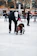 A person wearing a white cap and black clothing is helping a child in a red helmet who appears to be learning how to ice skate. The scene takes place on an ice rink with other skaters in the background and trees with no leaves. Orange cones are set up on the ice, possibly for guidance or lessons.