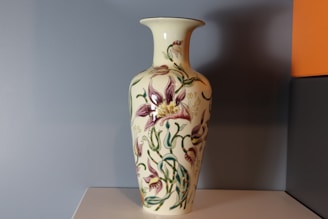 A decorative ceramic vase adorned with intricate floral patterns, featuring predominantly purple and green flowers on a cream background. It stands on a plain surface against a minimalistic, two-toned wall with muted gray and a hint of orange.