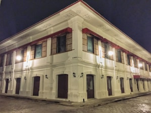 A large two-story colonial-style building with white walls and red accents is illuminated at night. The structure features traditional wooden windows with intricate lattice patterns and is adorned with decorative moldings along the roofline. Street lamps cast a warm glow across the cobblestone street surrounding the building, adding to its historical ambiance.