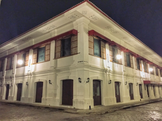 A large two-story colonial-style building with white walls and red accents is illuminated at night. The structure features traditional wooden windows with intricate lattice patterns and is adorned with decorative moldings along the roofline. Street lamps cast a warm glow across the cobblestone street surrounding the building, adding to its historical ambiance.
