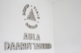 A white wall with silver or metallic text and a symbol. The symbol at the top consists of a series of geometric shapes surrounded by words. Below this, the text reads 'AULA' followed by 'DAARUT TAUHIID'.