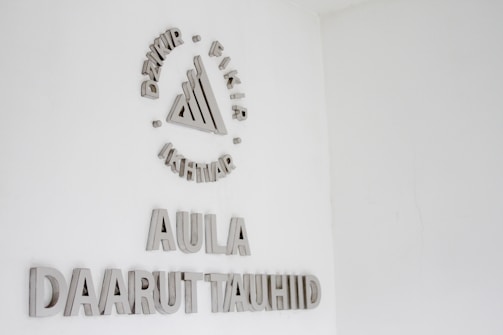 A white wall with silver or metallic text and a symbol. The symbol at the top consists of a series of geometric shapes surrounded by words. Below this, the text reads 'AULA' followed by 'DAARUT TAUHIID'.