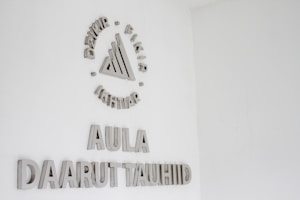 A white wall with silver or metallic text and a symbol. The symbol at the top consists of a series of geometric shapes surrounded by words. Below this, the text reads 'AULA' followed by 'DAARUT TAUHIID'.