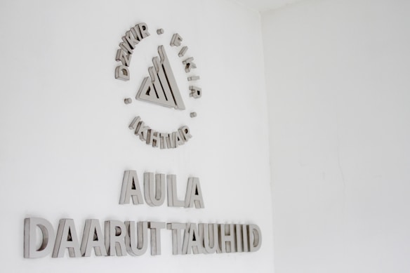 A white wall with silver or metallic text and a symbol. The symbol at the top consists of a series of geometric shapes surrounded by words. Below this, the text reads 'AULA' followed by 'DAARUT TAUHIID'.