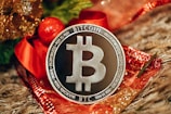 A silver Bitcoin coin is prominently displayed on a decorative background with red and gold accents, likely festive ribbons or ornaments. The coin's design includes the Bitcoin logo and inscriptions related to blockchain technology.