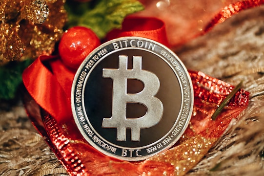 A silver Bitcoin coin is prominently displayed on a decorative background with red and gold accents, likely festive ribbons or ornaments. The coin's design includes the Bitcoin logo and inscriptions related to blockchain technology.