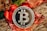 A silver Bitcoin coin is prominently displayed on a decorative background with red and gold accents, likely festive ribbons or ornaments. The coin's design includes the Bitcoin logo and inscriptions related to blockchain technology.