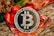 A silver Bitcoin coin is prominently displayed on a decorative background with red and gold accents, likely festive ribbons or ornaments. The coin's design includes the Bitcoin logo and inscriptions related to blockchain technology.