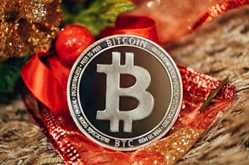 A silver Bitcoin coin is prominently displayed on a decorative background with red and gold accents, likely festive ribbons or ornaments. The coin's design includes the Bitcoin logo and inscriptions related to blockchain technology.