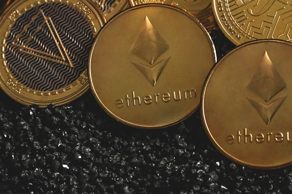 Several cryptocurrency coins, prominently displaying the Ethereum symbol and name, rest closely together. The background features a textured, black granular surface, creating a contrasting backdrop for the shiny, gold-colored coins.