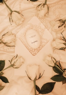 An arrangement features a white Chanel hand cream product placed on a white fabric amidst scattered pearl beads and several white roses. The elegant composition suggests a luxurious and refined aesthetic.